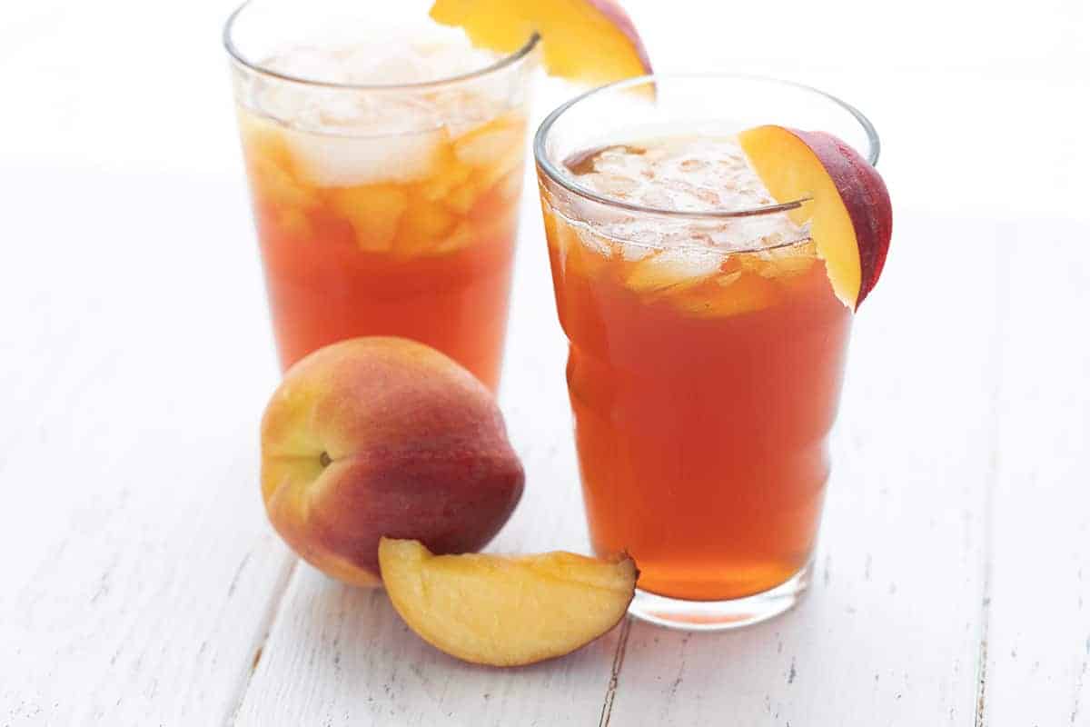 Två glas söt persika te på ett vitt bord med skivade persikor.