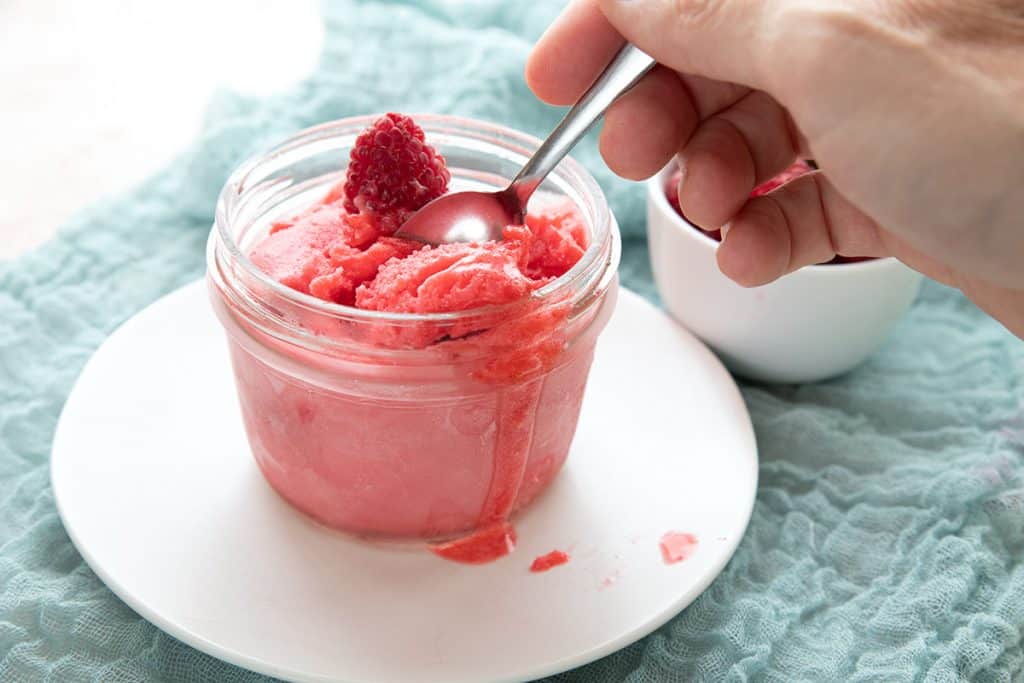 A hand digging into a jar of keto raspberry sorbet with a small spoon.