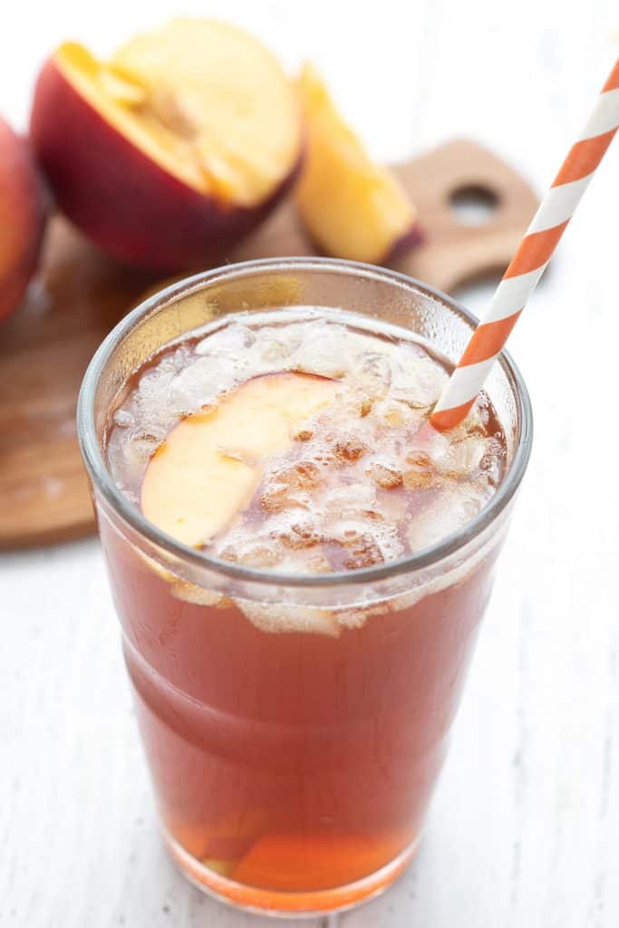 A glass filled with peach keto sweet tea, with a cutting board filled with cut peaches in the background.