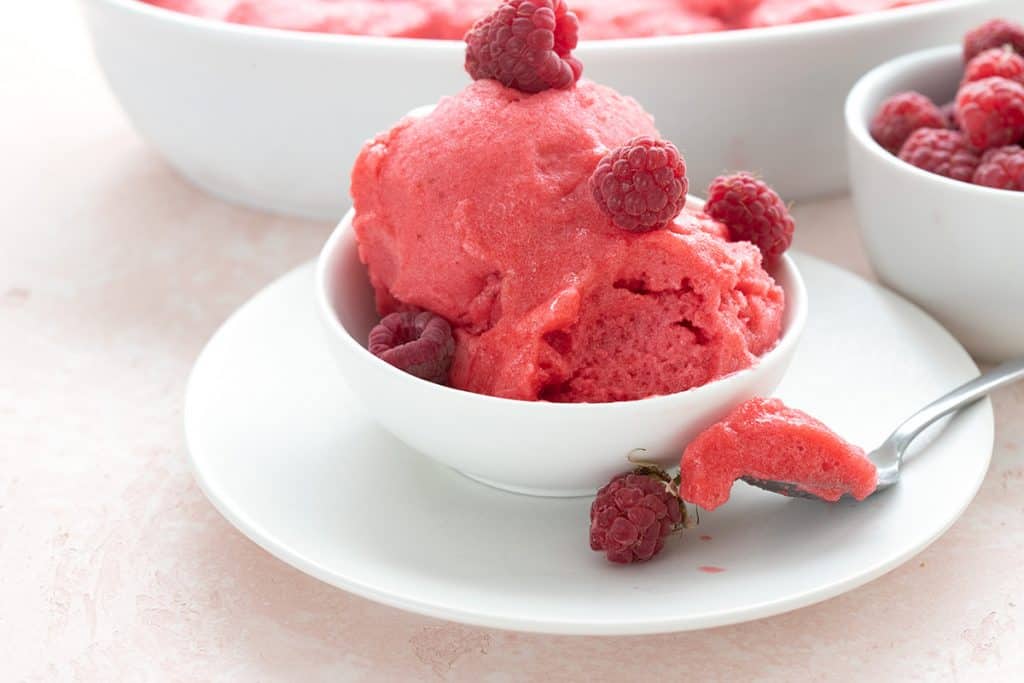 Sugar free raspberry sorbet in a white bowl on a white plate, with a spoonful taken out.