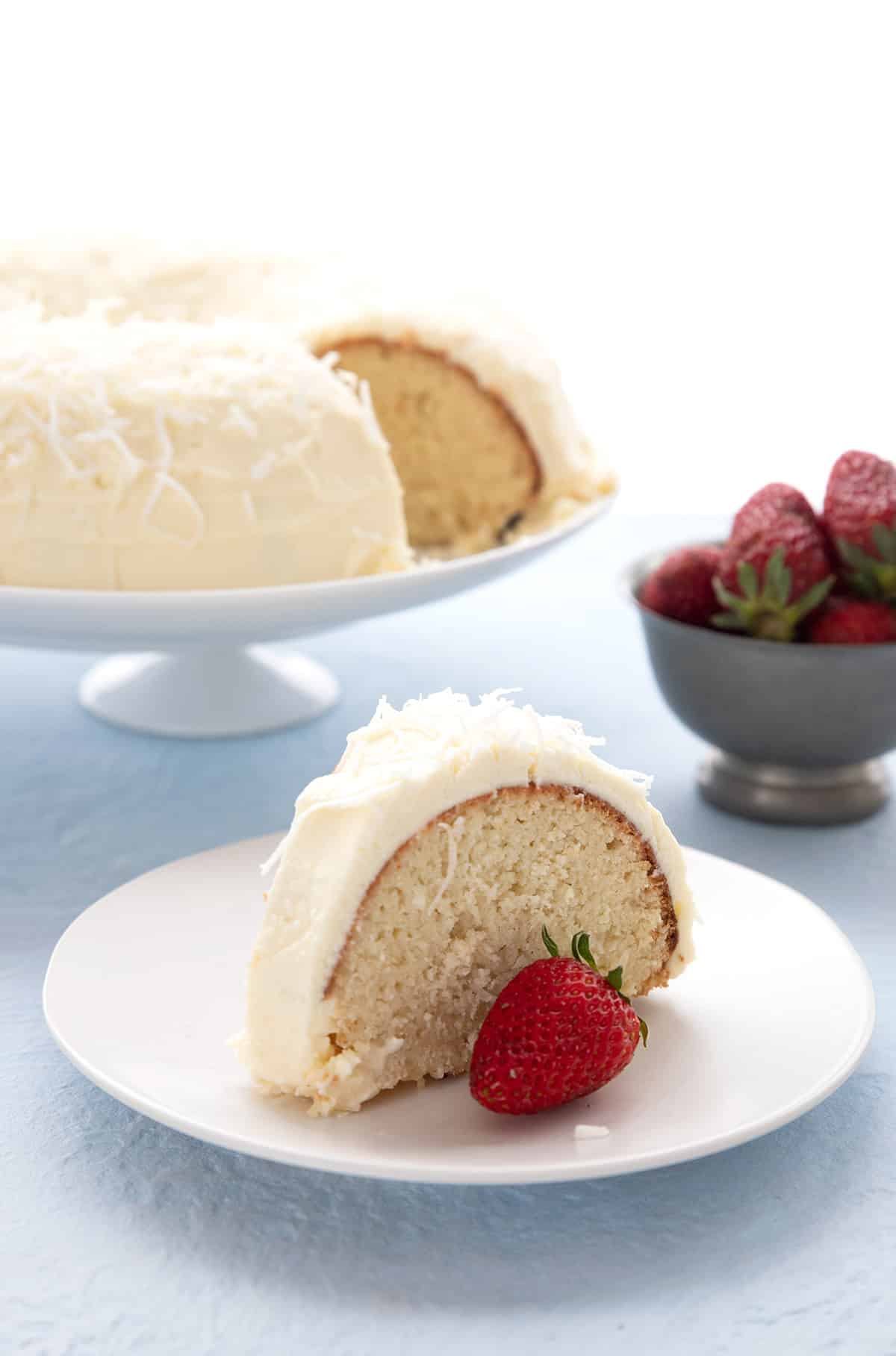 A slice of keto coconut cake on a white plate with a bowl of strawberries and the rest of the cake in the background.