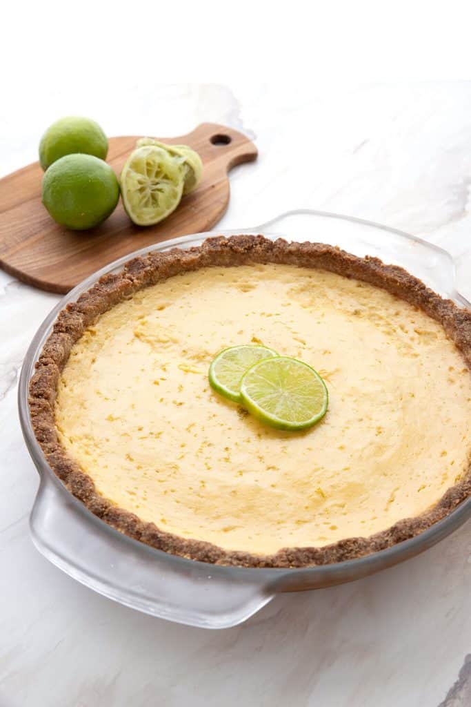 Keto key lime pie in a glass pie dish in front of a wooden cutting board with limes on it.