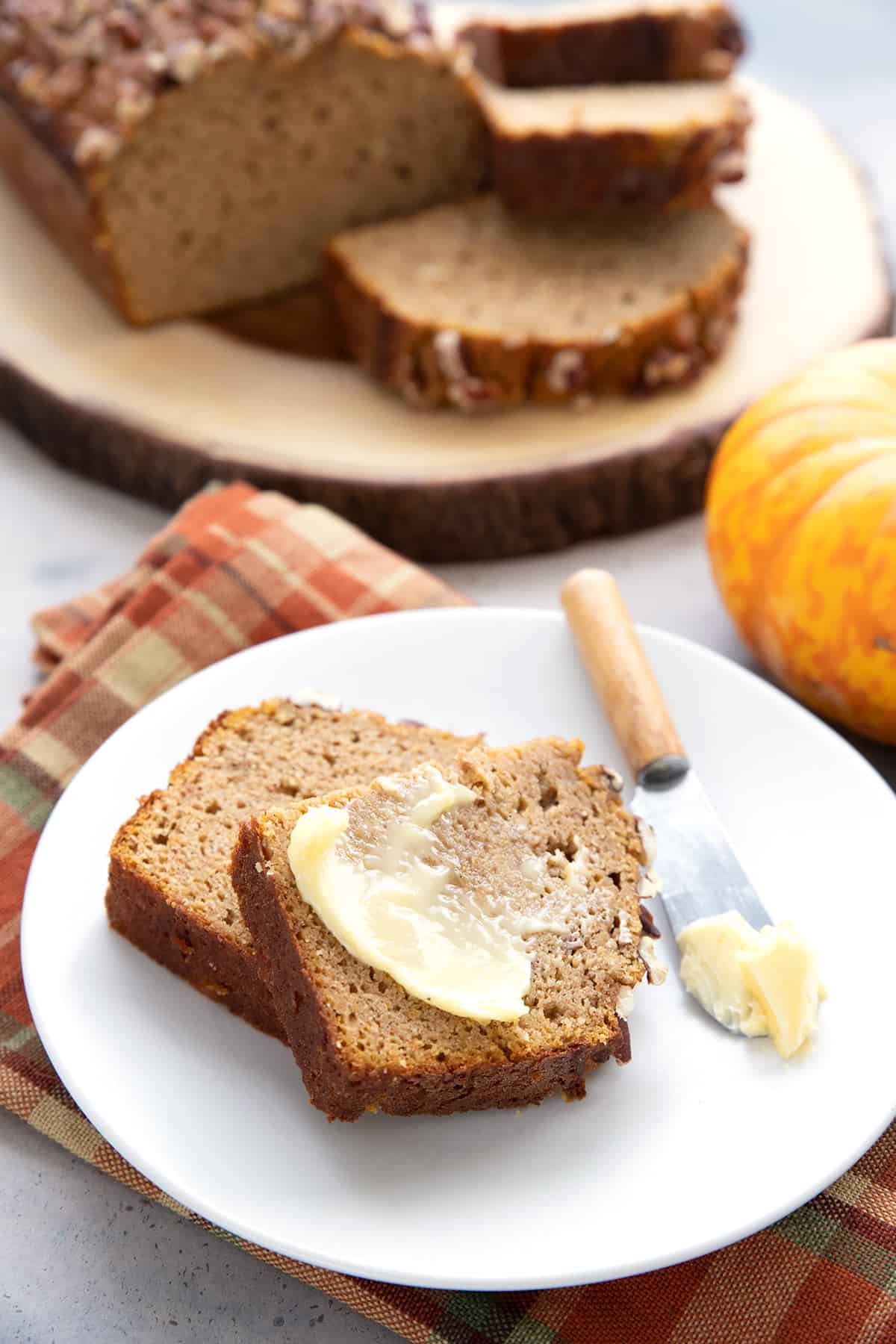 A slice of keto pumpkin bread with butter on it, on a white plate over a plaid napkin.