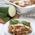 A slice of zucchini lasagna on a white plate in front of the pan, with some zucchini on a wooden cutting board.