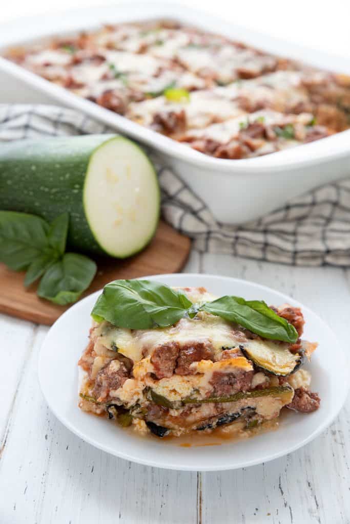 A slice of zucchini lasagna on a white plate in front of the pan, with some zucchini on a wooden cutting board.