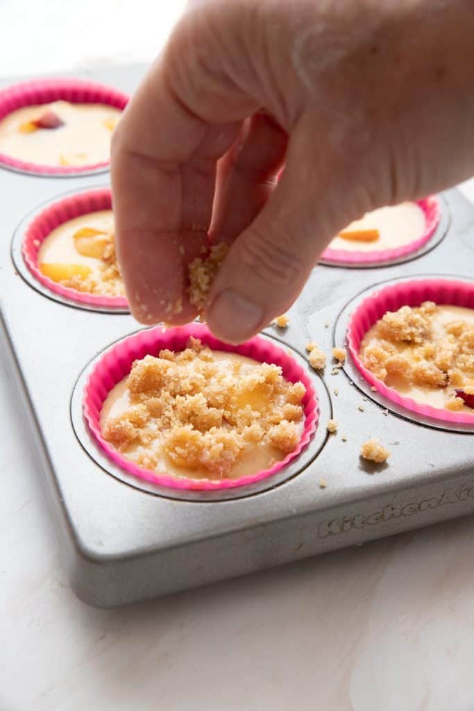 A hand sprinkling streusel over peach muffins before baking.