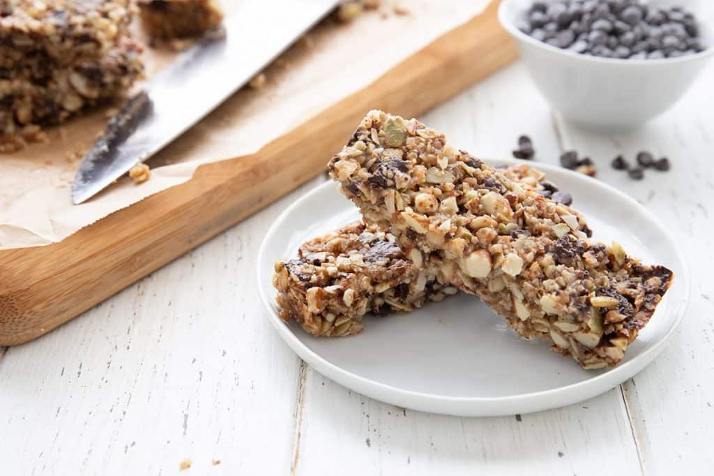 Two grain free keto granola bars on a white plate in front of a bowl of mini sugar free chocolate chips.