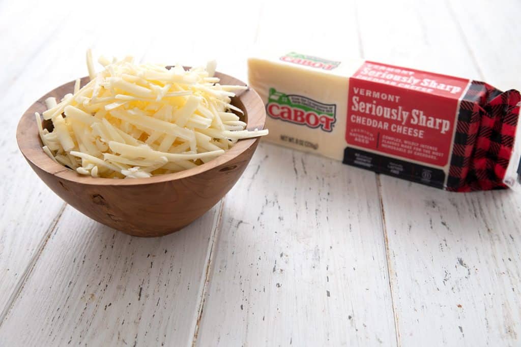 A wooden bowl of shredded cheese beside a block of cheddar.