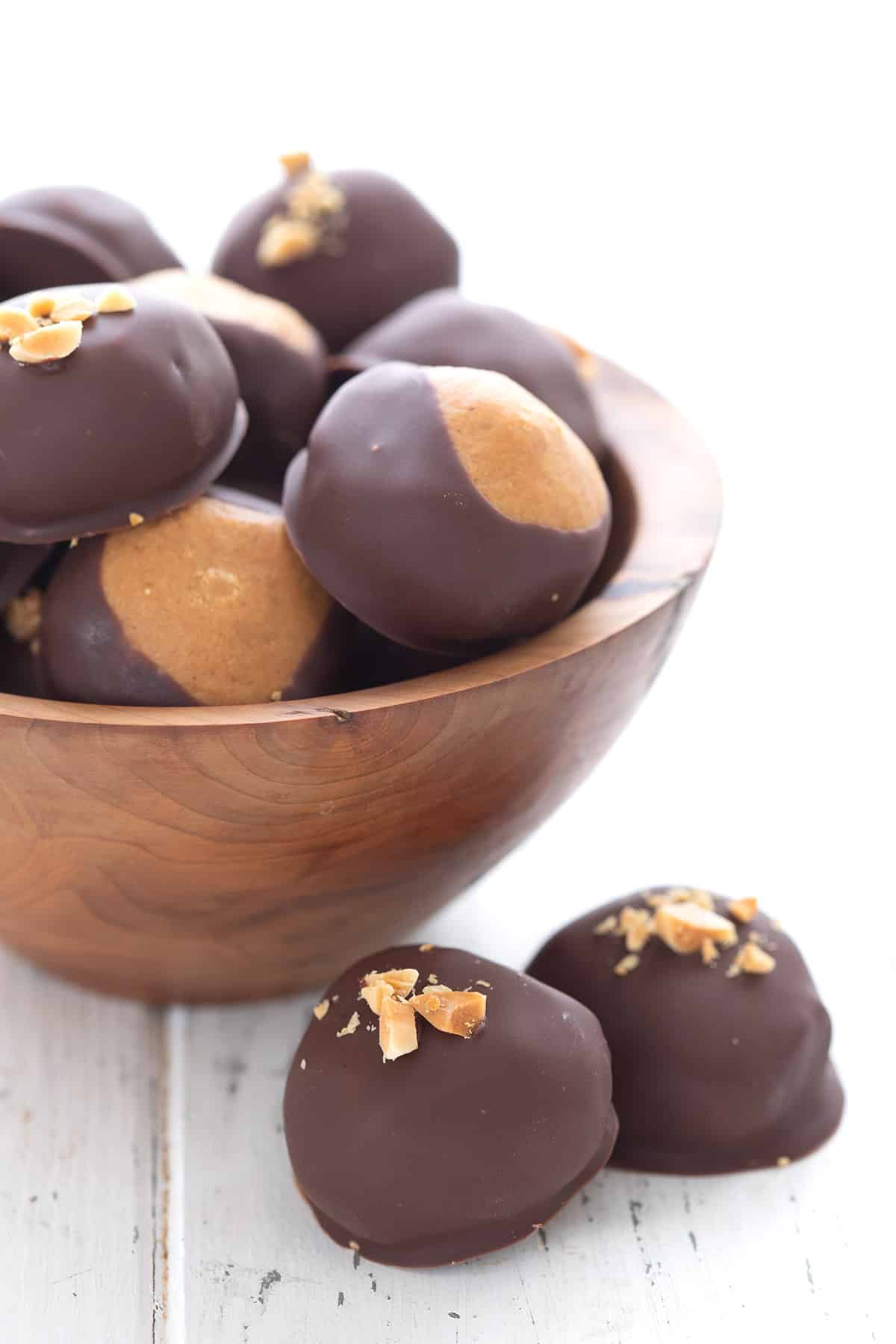 Keto peanut butter balls on a white table in front of a wooden bowl holding more balls.