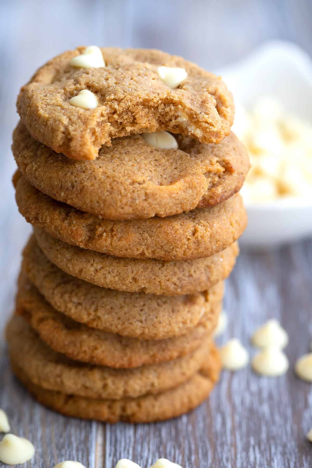 A stack of keto pumpkin cookies with white chocolate chips on a wooden table with a bite taken out of the top one.