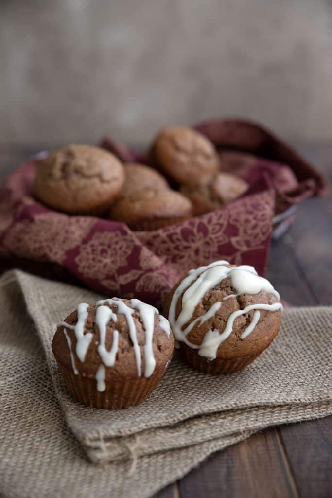 Two glazed keto gingerbread muffins on a burlap napkin in front of a bowl of more muffins.