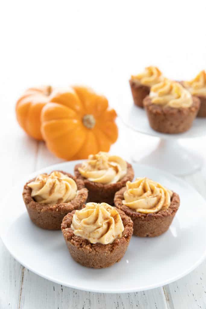 Four keto pumpkin cheesecake bites on a white plate with mini pumpkins in the background.