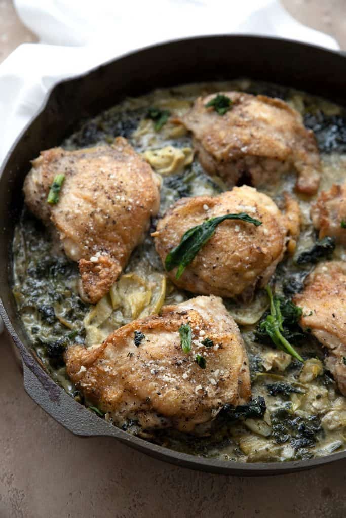Six crispy chicken thighs with spinach and artichokes in a cast iron skillet.