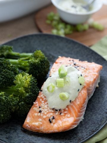 A piece of roasted keto salmon on a black plate with wasabi mayonnaise and chopped green onions on top.