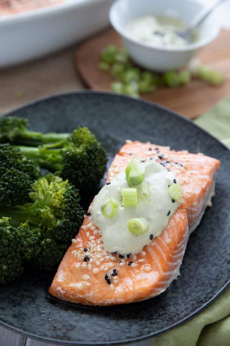 A piece of roasted keto salmon on a black plate with wasabi mayonnaise and chopped green onions on top.