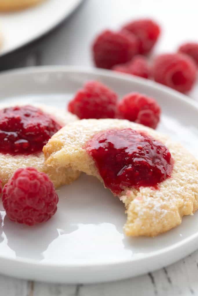 Close up shot of a keto jam thumbprint cookie on a white plate with raspberries in the background.