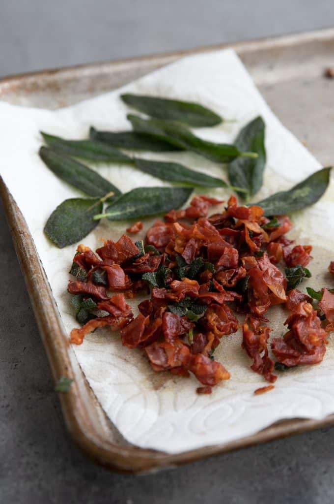 Crispy prosciutto and sage leaves draining on a white paper towel.