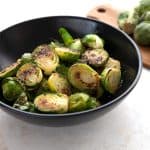 Caramelized Brussels sprouts in a black bowl with a cutting board full of sprouts in the background.
