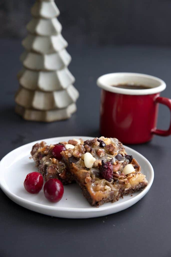 Two keto cranberry pecan magic bars on a small white plate in front of a red cup of coffee.