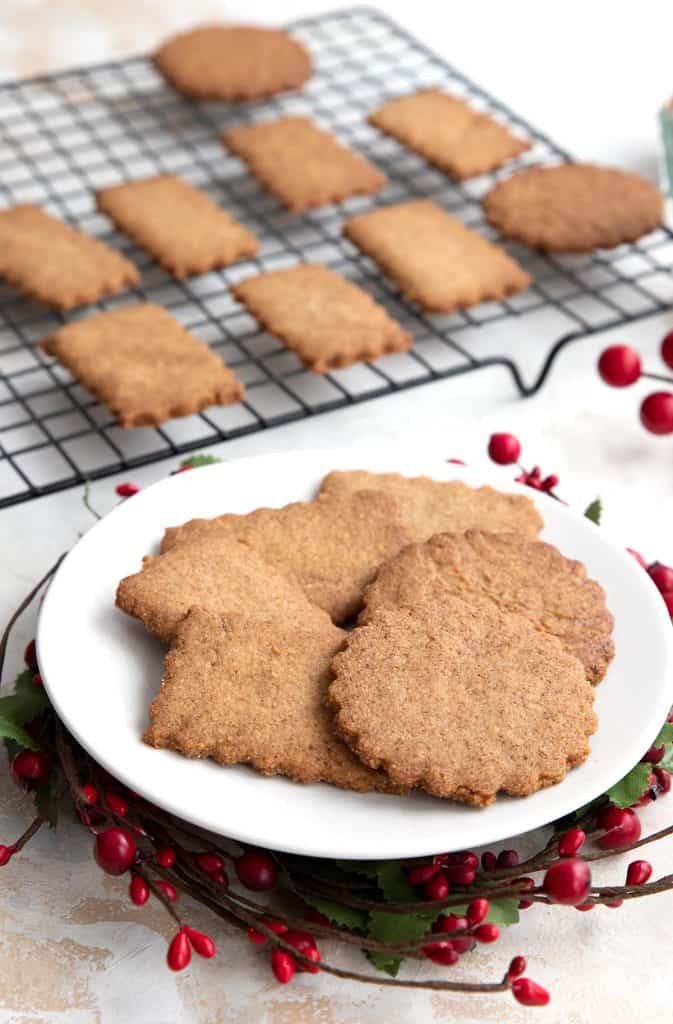 Keto Biscoff cookies sit on a white plate over a wreath of holly berries, with more cookies in the background.