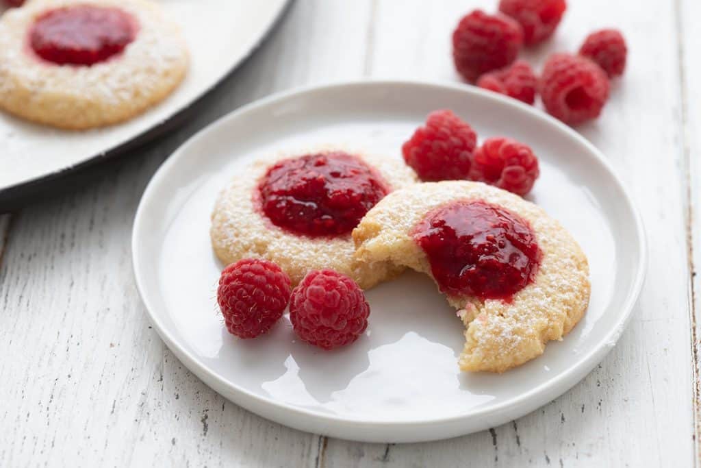 Two keto thumbprint cookies on a white plate with raspberries all around.