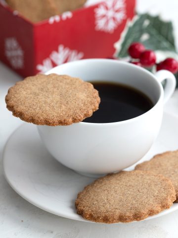 A keto speculoos cookie sits on the edge of a cup of coffee, with more cookies sitting in the saucer.