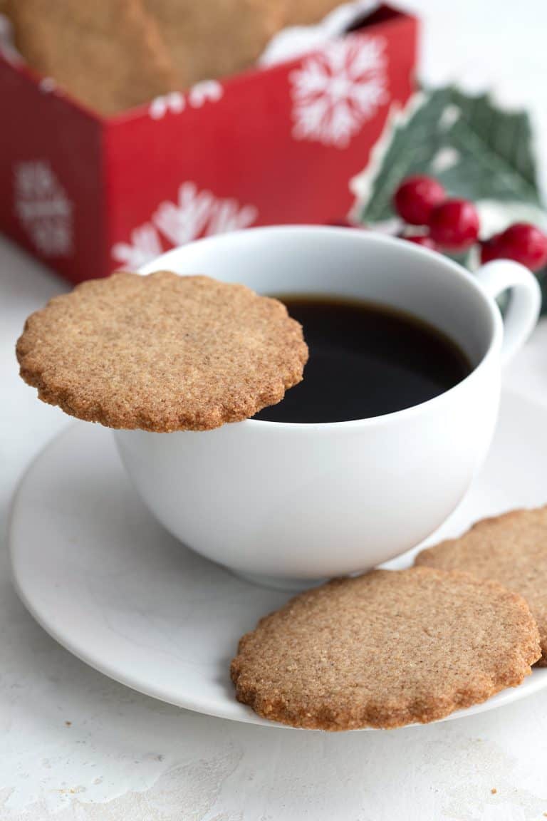 A keto speculoos cookie sits on the edge of a cup of coffee, with more cookies sitting in the saucer.