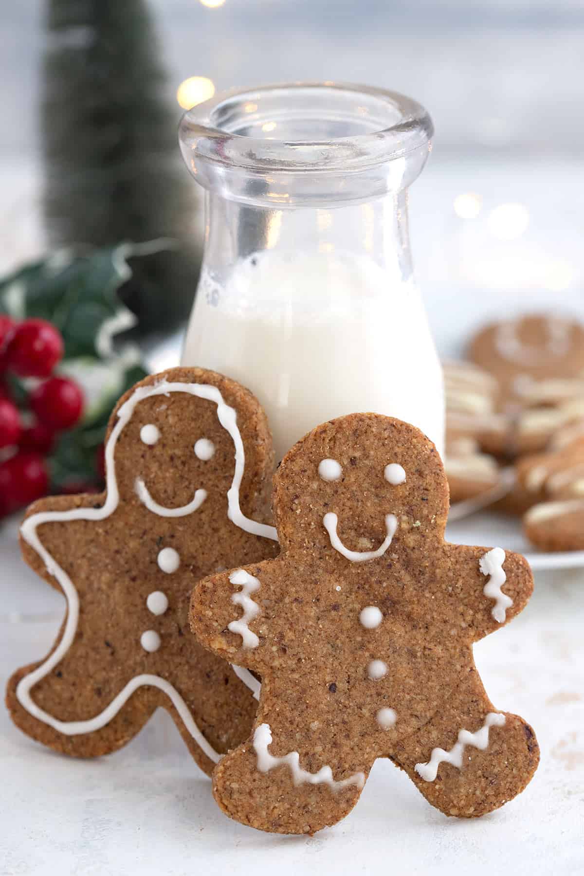 Two keto gingerbread cookies leaning up against a glass bottle of milk, with more cookies and Christmas decorations in the background.