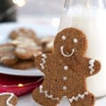 A keto gingerbread man leaning up against a glass of milk, with a plate of cookies in the background.
