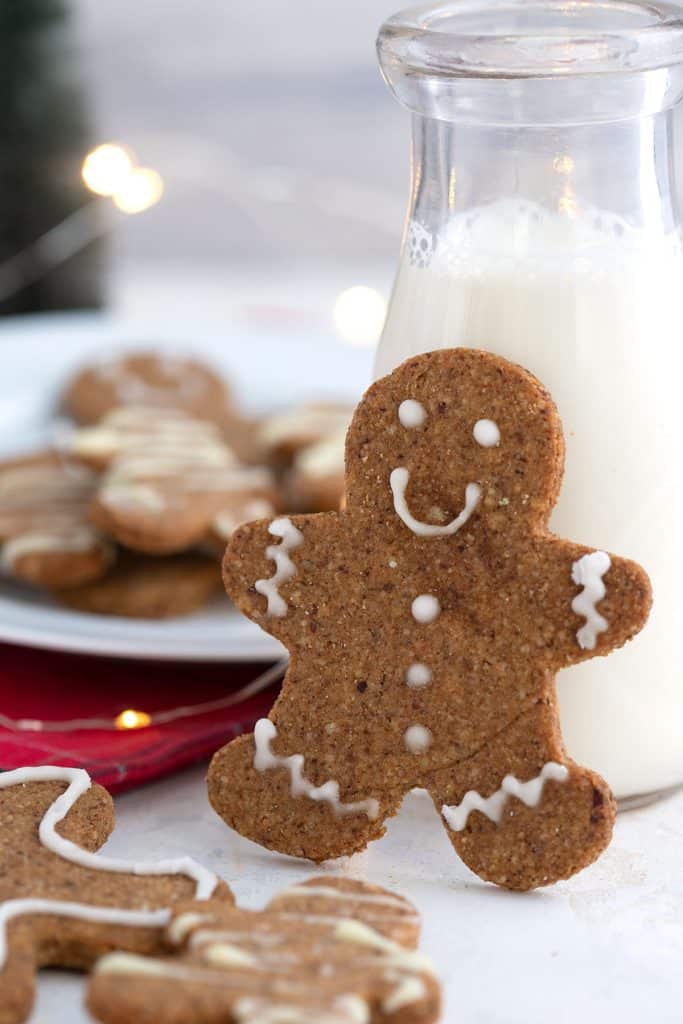 A keto gingerbread man leaning up against a glass of milk, with a plate of cookies in the background.