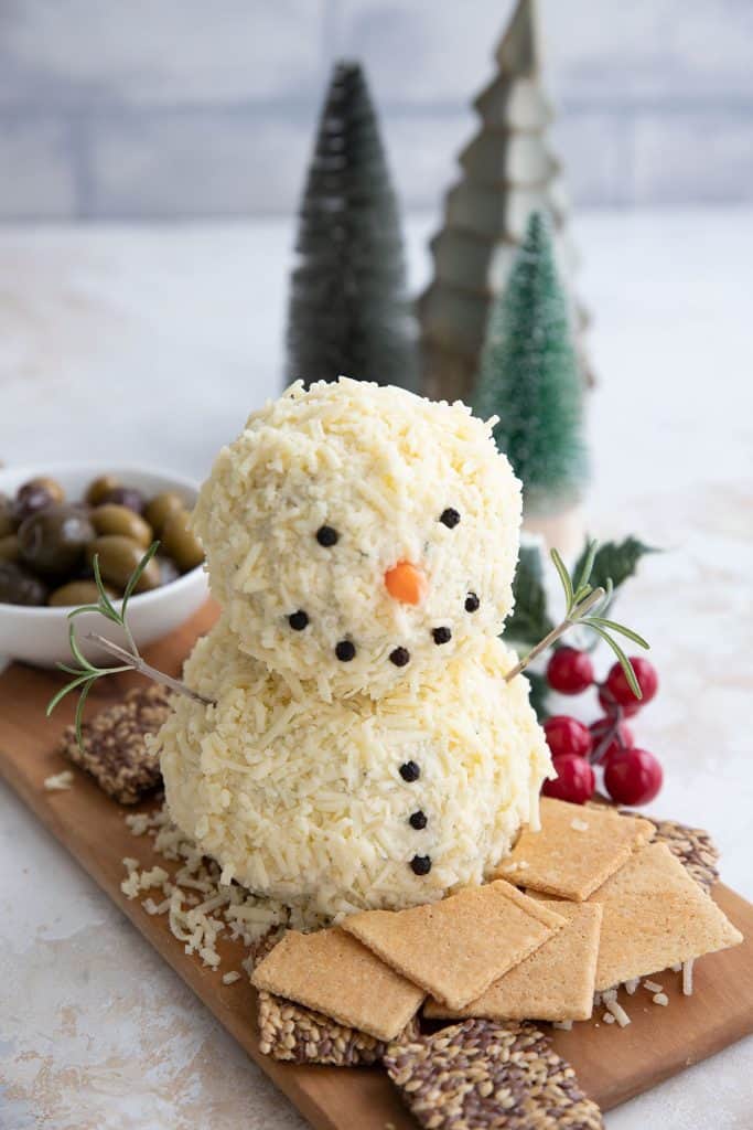 A snowman cheeseball on a wooden cutting board, surrounded by keto crackers, with Christmas decorations in the background.