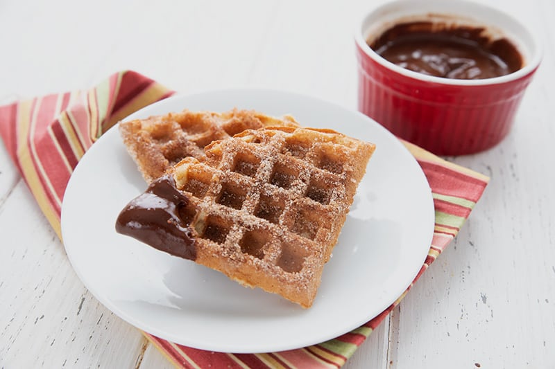 Two keto churro waffles on a white plate, with chocolate sauce in a pot beside the plate. 