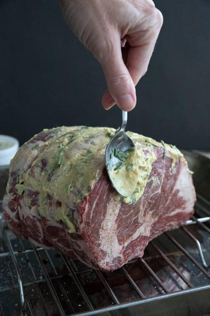 Slathering Dijon and herbs over a standing rib roast in a roasting pan.
