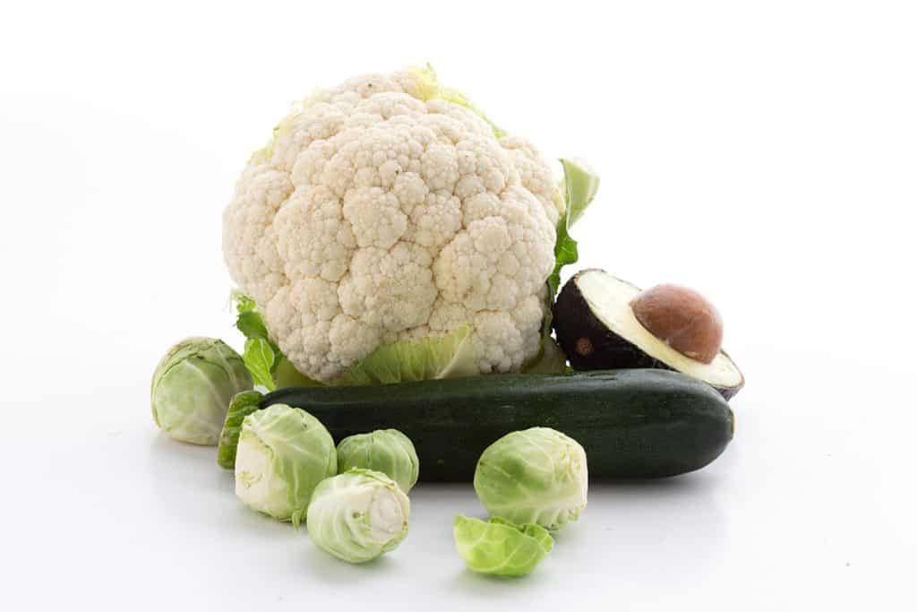 A heat of cauliflower, a zucchini, brussels sprouts and half an avocado on a white background.