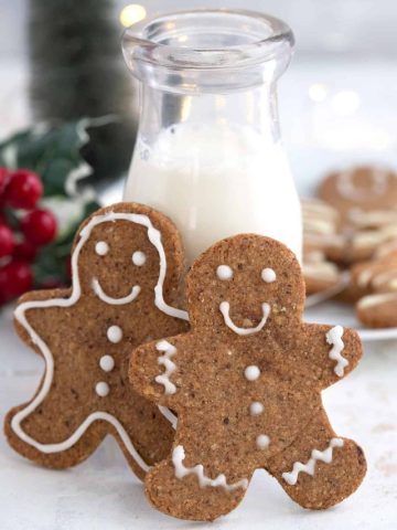 Two keto gingerbread cookies leaning up against a glass bottle of milk, with more cookies and Christmas decorations in the background.
