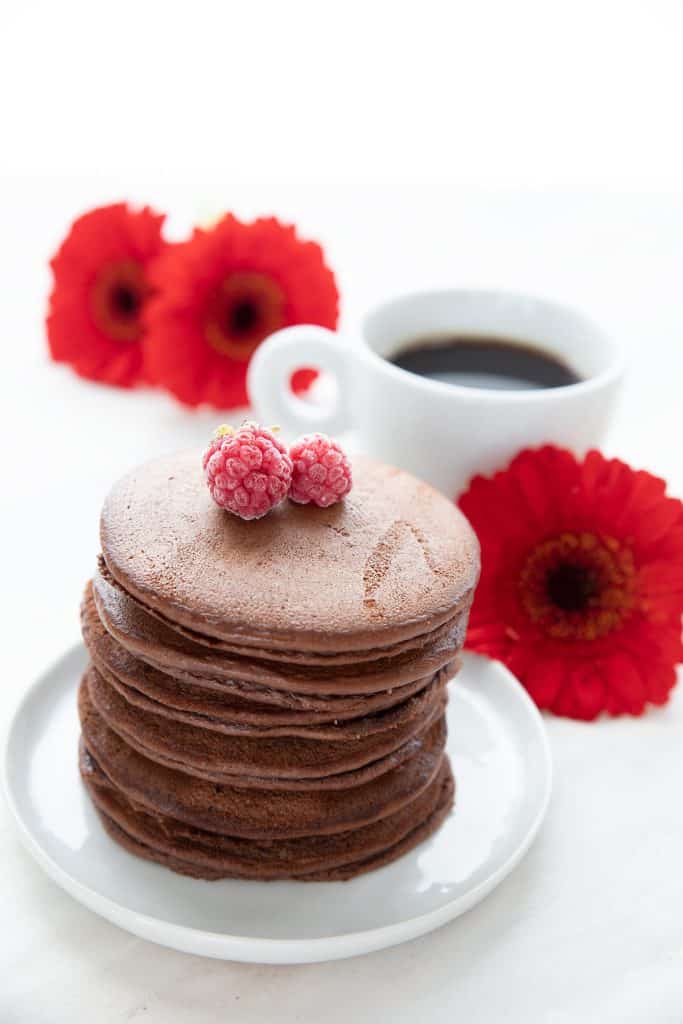 A stack of chocolate protein pancakes on a white plate, with red Gerbera daisies and a cup of coffee in the background.