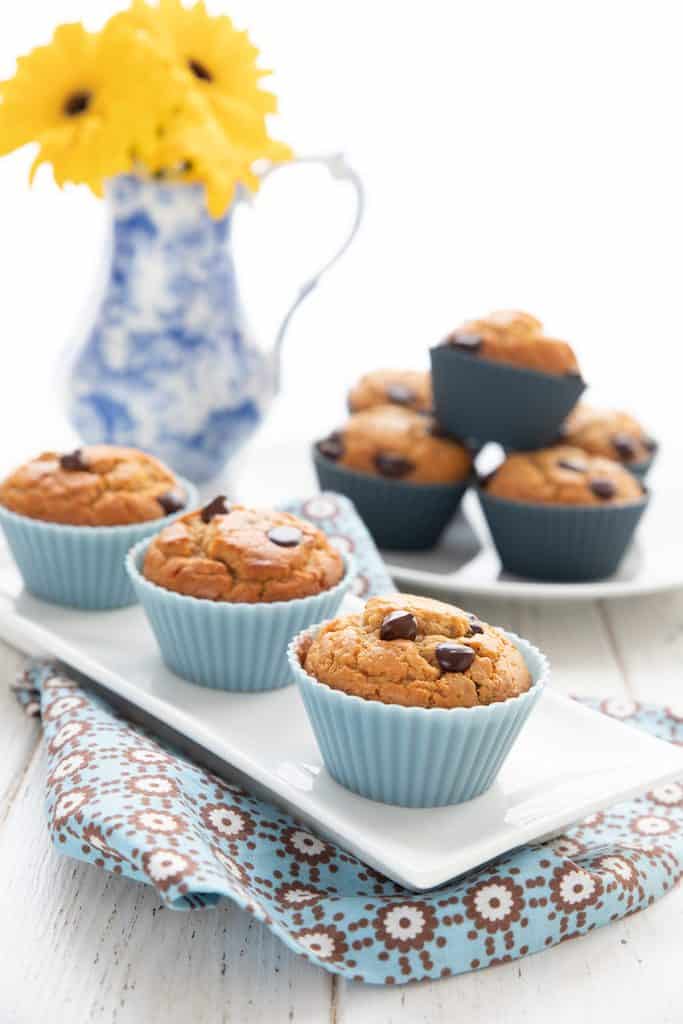 Three keto peanut butter muffins in blue liners on a white platter, in front of a vase with flowers and another plate of muffins.