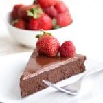 A slice of keto flourless chocolate cake on a white plate in front of a bowl of berries.