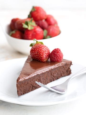 A slice of keto flourless chocolate cake on a white plate in front of a bowl of berries.