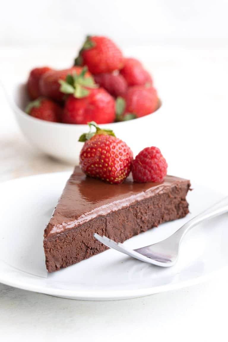 A slice of keto flourless chocolate cake on a white plate in front of a bowl of berries.