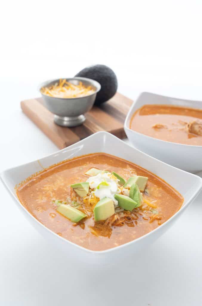 Two bowls of easy chicken enchilada soup with cheese and an avocado in the background.