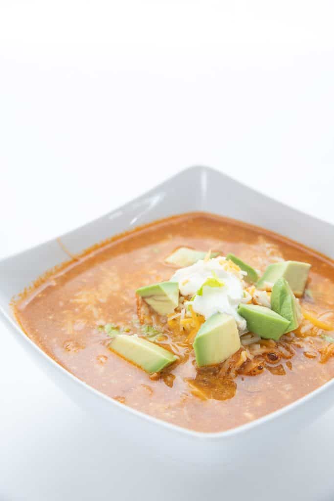 A square white bowl full of keto chicken enchilada soup with sour cream and chopped avocado on top.