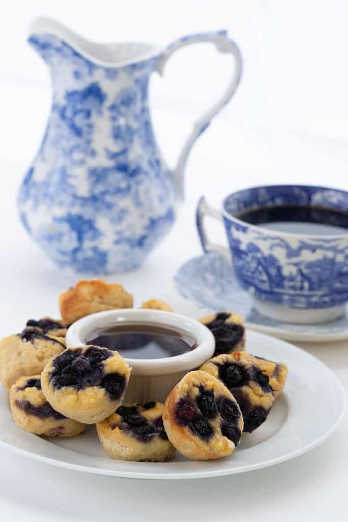 Keto blueberry pancakes bites on a white plate, with a cup of coffee and a blue vase in the background.