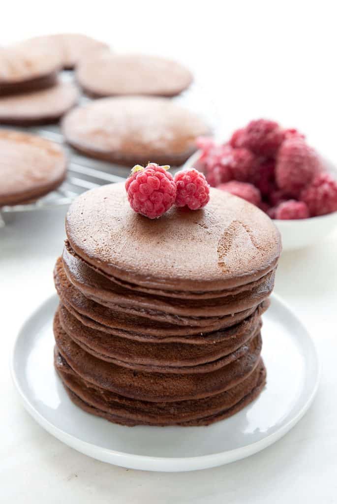 a stack of chocolate protein pancakes on a white plate with raspberries on top.