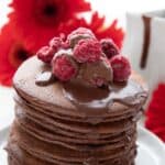 A stack of low carb chocolate protein pancakes on a white plate, with raspberries and chocolate sauce on top.