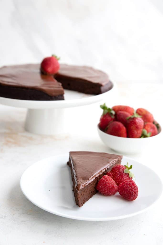 A slice of flourless chocolate cake on a white plate with berries, with the rest of the cake in the background.