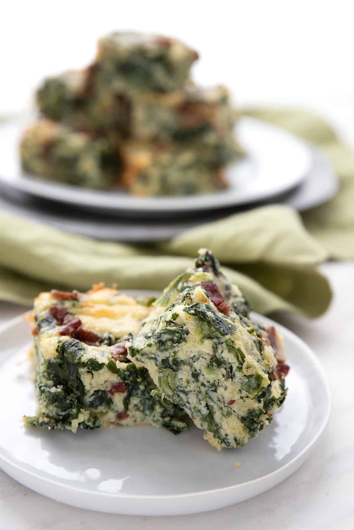 Two cheesy spinach squares on a white plate in front of a platter of more squares.