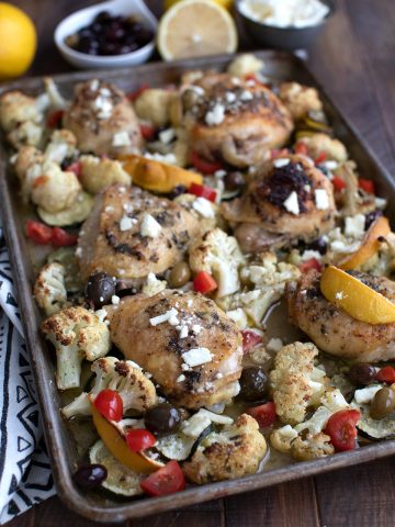 A sheet pan full of Greek Lemon Chicken with vegetables, and lemon and olives in the background.