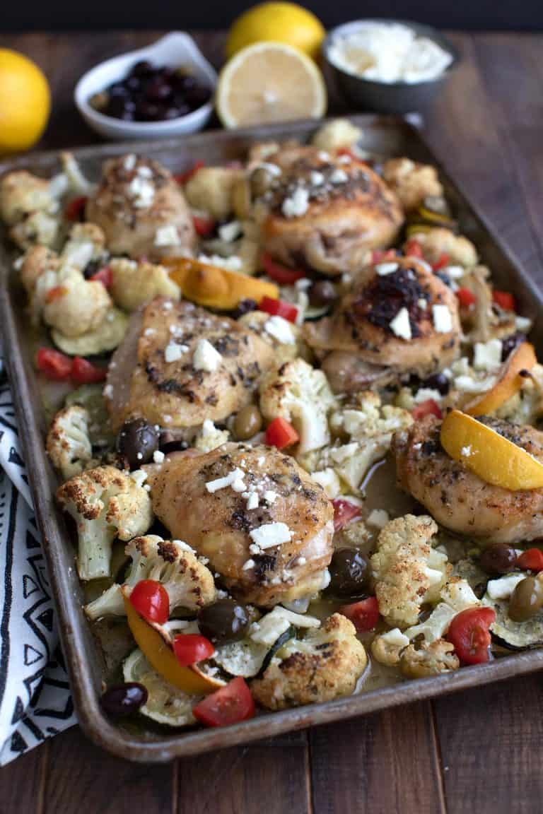 A sheet pan full of Greek Lemon Chicken with vegetables, and lemon and olives in the background.