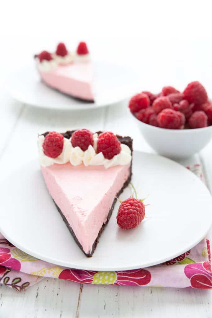 Two slices of keto raspberry mousse tart on white plates with a bowl of raspberries.